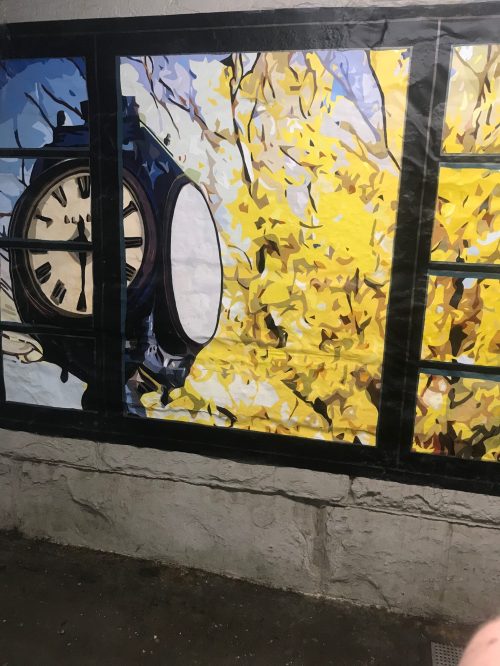 A mural featuring a clock and vibrant yellow leaves, framed by a window-like design on a wall.