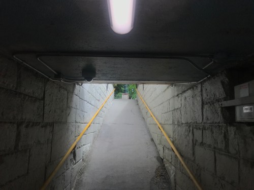A dimly lit tunnel with a concrete ceiling and yellow handrails leading to a bright exit at the end.