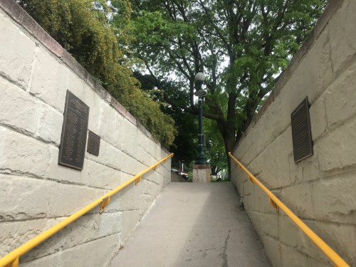 A pathway with yellow railings leads up between stone walls, flanked by plaques, under a green tree canopy.