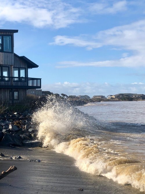 Waves crash onto a sandy shore near a wooden house, with a cloudy blue sky in the background.