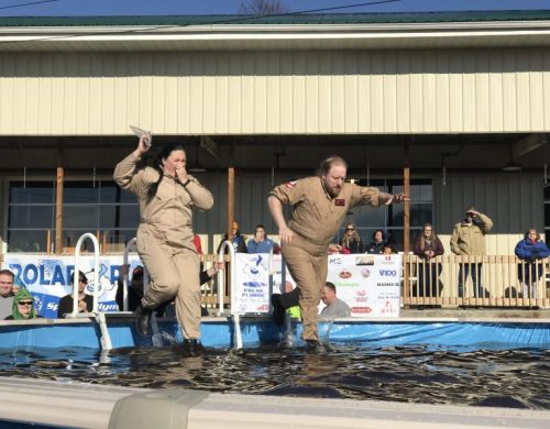 Two people in beige jumpsuits are jumping into a pool, with a crowd watching from the sidelines.