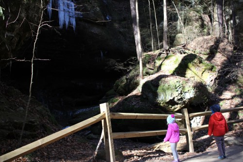 Two children stand near a wooden fence, looking at a rocky area with icicles in a forested setting.