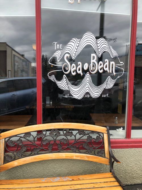 A wooden bench in front of a café window displaying the logo "The Sea Bean" with a wavy design.