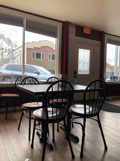 A cozy café interior with wooden tables and black chairs, featuring large windows and a view of the street outside.
