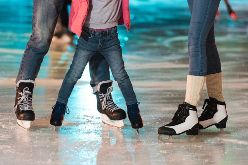 A child in jeans and a pink jacket skates between two adults on an ice rink, all wearing ice skates.