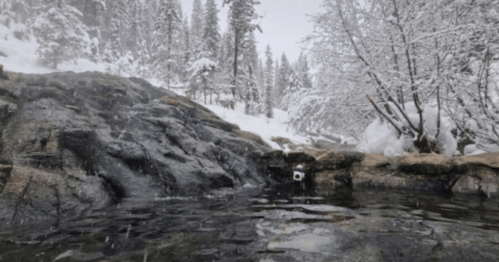 A snowy landscape with a rocky shoreline and a calm water pool, surrounded by trees and falling snowflakes.