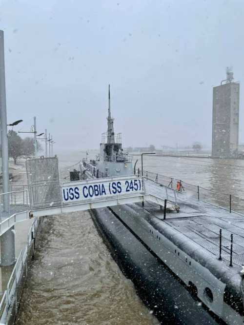 A submarine, USS Cobia (SS 245), docked in snowy weather with a river and industrial structures in the background.