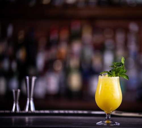 A refreshing yellow cocktail garnished with mint, set against a blurred bar background.