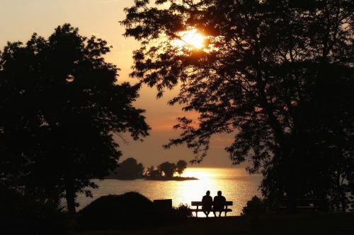 Silhouetted couple sitting on a bench by the water, watching a sunset through trees.