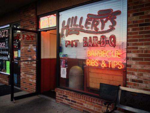 Exterior of Hillery's Pit Bar-B-Q with neon signs advertising barbecue, ribs, and tips, and an open door.