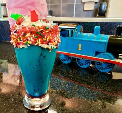 A colorful blue milkshake topped with whipped cream, sprinkles, and cotton candy, next to a toy train.
