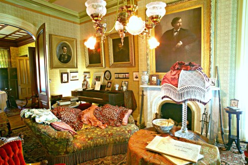 A vintage parlor with ornate decor, featuring a piano, portraits, a lamp, and a richly patterned sofa.