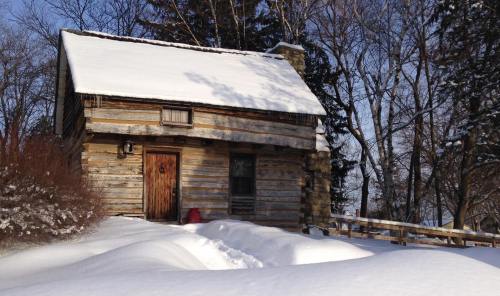 A rustic wooden cabin covered in snow, surrounded by trees and a snowy landscape under a clear blue sky.
