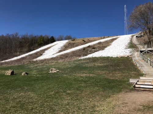 A grassy hill with two snow-covered slopes under a clear blue sky, featuring a wooden staircase on the right.