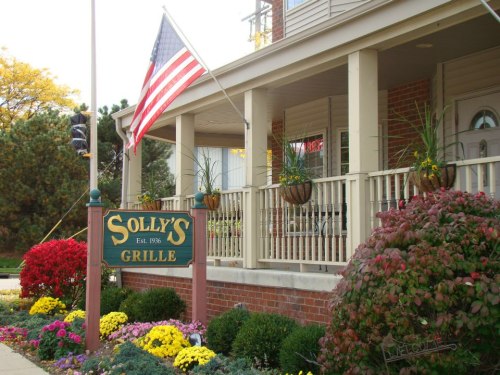 Exterior of Solly's Grille with an American flag, colorful flowers, and a welcoming porch. Established in 1996.