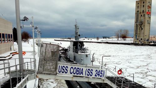 The USS Cobia (SS 245) submarine docked in a snowy harbor, surrounded by ice and overcast skies.