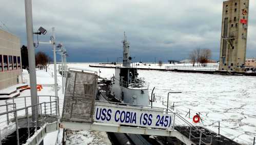 The USS Cobia (SS 245) submarine docked in a snowy harbor, surrounded by ice and overcast skies.