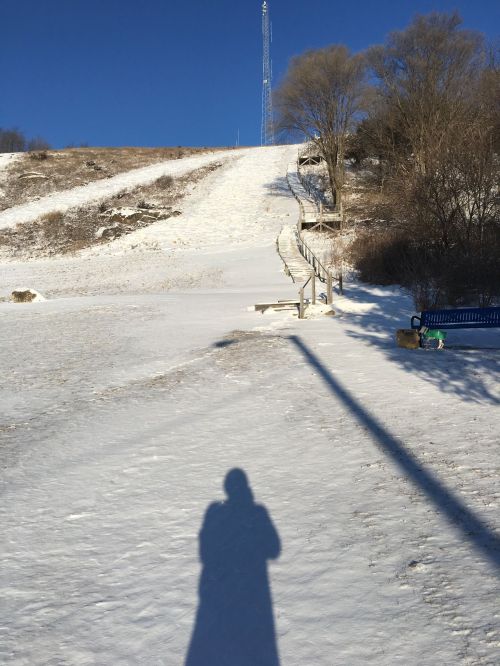 A snowy hill with a clear blue sky, featuring a shadow in the foreground and a bench nearby.
