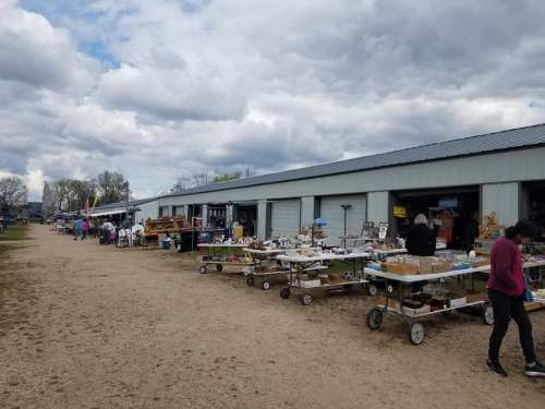 A bustling outdoor market with vendors displaying various items on tables and carts under a cloudy sky.