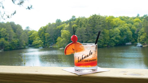 A refreshing cocktail with a cherry and orange slice sits on a wooden railing, overlooking a serene lake and lush trees.