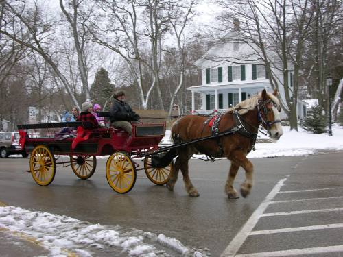 A horse-drawn carriage with passengers rides through a snowy street, passing a white house with green shutters.