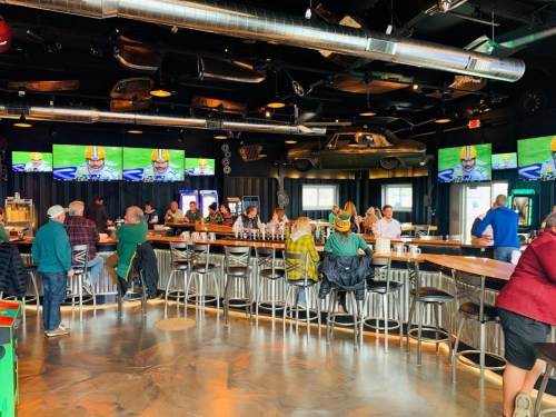 A lively bar scene with patrons enjoying drinks, large screens showing a sports game, and a modern industrial decor.