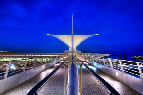 A modern architectural structure with a pointed canopy, illuminated against a deep blue evening sky.