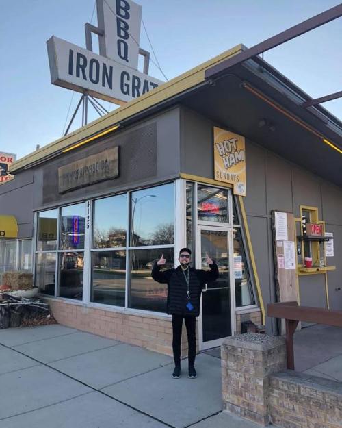 A person stands outside Iron Grate BBQ, giving a thumbs up in front of the restaurant's sign and entrance.