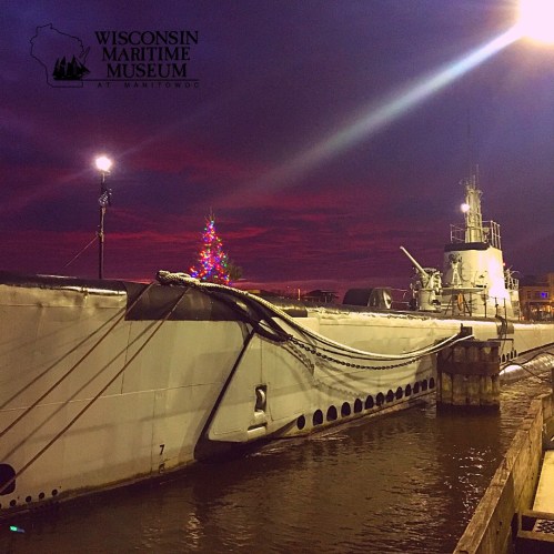 A submarine docked at night, illuminated by colorful lights, with a festive tree and a sunset in the background.