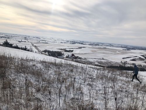 A person walks on a snowy hillside, overlooking a vast landscape of fields and distant hills under a cloudy sky.