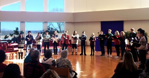 A choir performs in a bright room with large windows, surrounded by an audience seated in chairs.