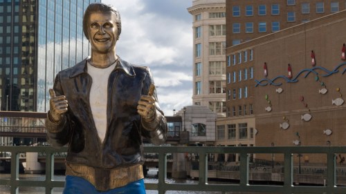 A bronze statue of a smiling man in a leather jacket, standing by a river with buildings in the background.