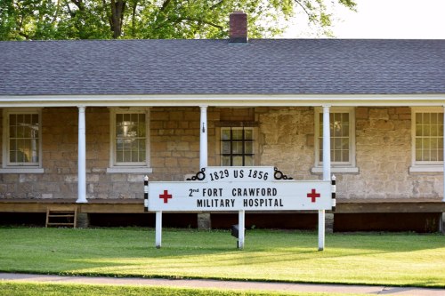 Historic building with a sign reading "2nd Fort Crawford Military Hospital," established 1829, US 1856. Green lawn in front.