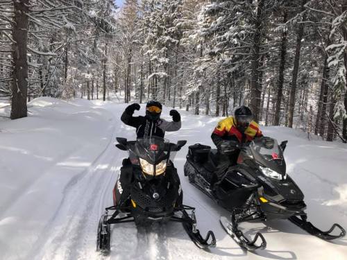 Two snowmobilers pose in a snowy forest, surrounded by tall trees, with one flexing their muscles.