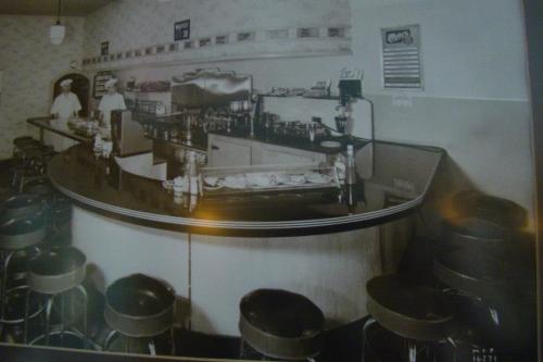Black and white photo of a vintage diner interior with a curved counter and bar stools, featuring two staff members.