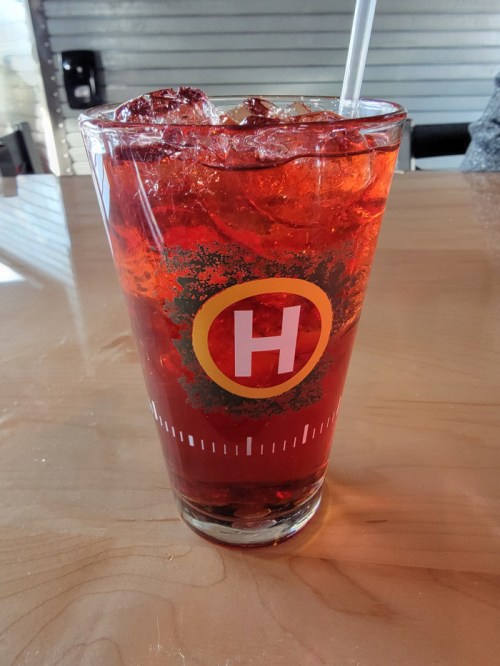 A tall glass filled with a red beverage and ice, featuring a logo with a yellow circle and the letter "H."
