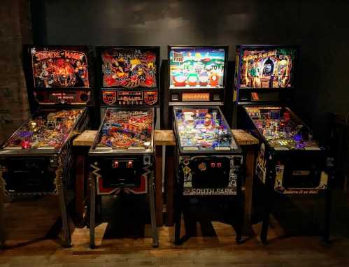 A row of five colorful pinball machines in a dimly lit arcade setting.