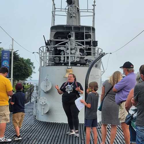 A guide speaks to a group of visitors near a historic submarine on a dock, with trees and water in the background.