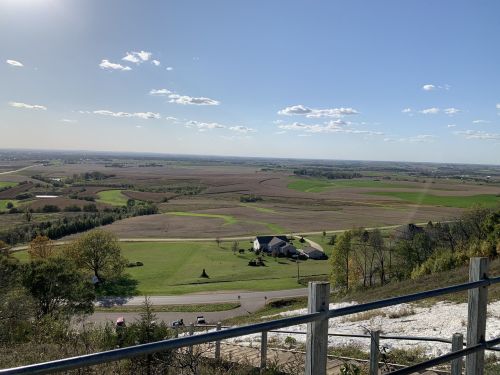 A panoramic view of rolling fields and a distant house under a clear blue sky with scattered clouds.