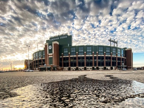 Lambeau Field under a dramatic sky, reflecting puddles on the ground, with the sun setting in the background.