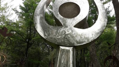 A large metallic sculpture resembling a female symbol, surrounded by trees in a natural setting.