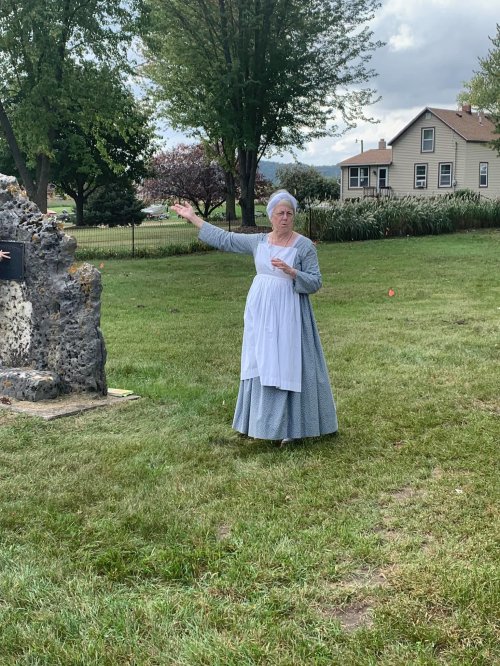 A woman in historical attire gestures while standing near a stone structure in a grassy area with trees and houses in the background.