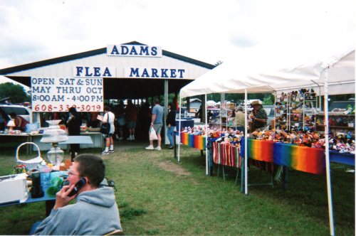 A bustling flea market with vendors under tents, colorful displays, and a sign for "Adams Flea Market" in the background.