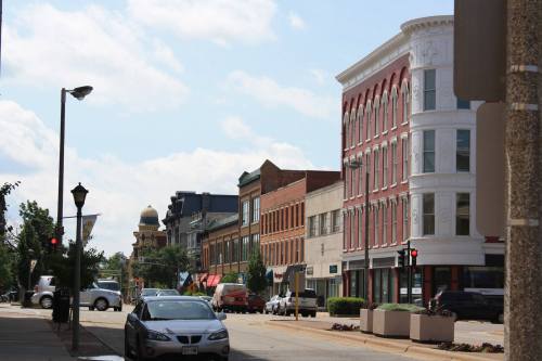 A street view of a small town with historic buildings, parked cars, and a partly cloudy sky.