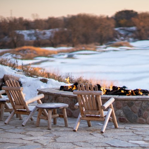 A cozy outdoor seating area with wooden chairs around a stone fire pit, overlooking a serene landscape at sunset.