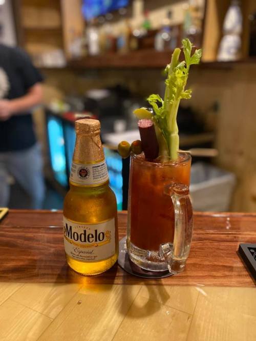 A bottle of Modelo beer next to a tall glass of a Bloody Mary garnished with celery and a sausage.