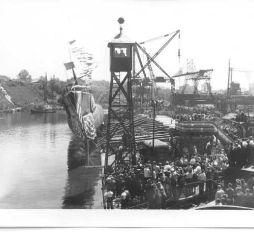 A crowd gathers by the water as a large ship is launched, with cranes and a watchtower in the background.