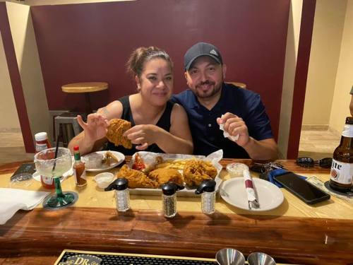 A couple enjoys a meal of fried chicken at a restaurant, smiling and holding food, with drinks and condiments on the table.