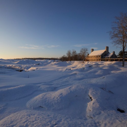 A snowy landscape with rolling hills and a rustic building in the background under a clear blue sky.