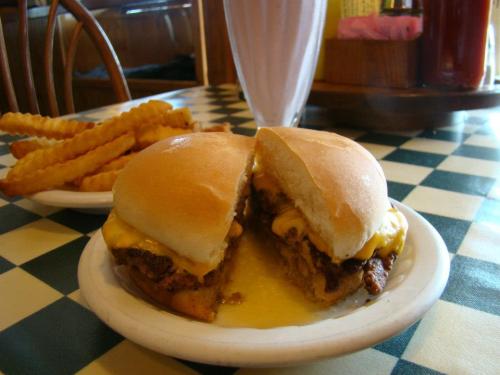 A plate with two cheeseburgers, melted cheese oozing out, alongside a serving of crinkle-cut fries and a milkshake.
